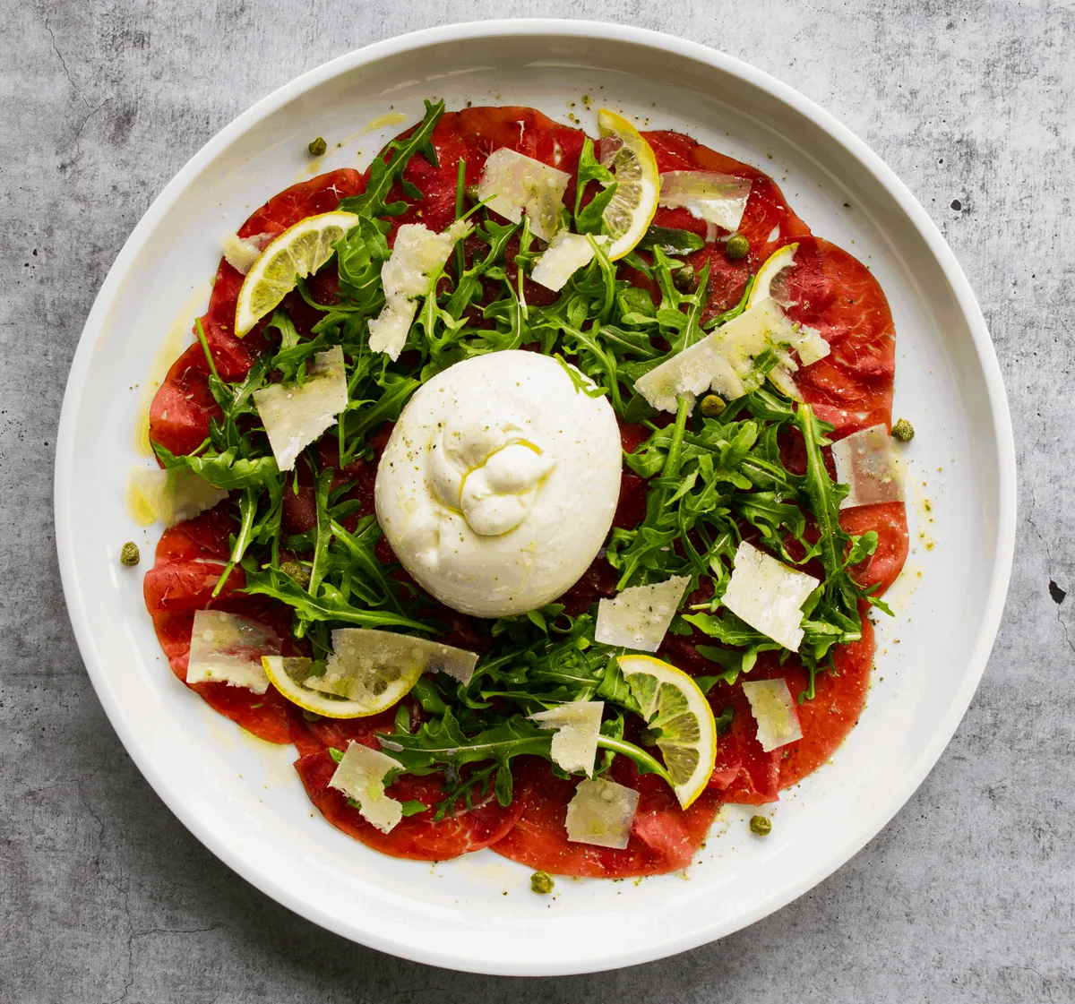 Thinly sliced cured meat with arugula, shaved cheese and lemon on a white plate